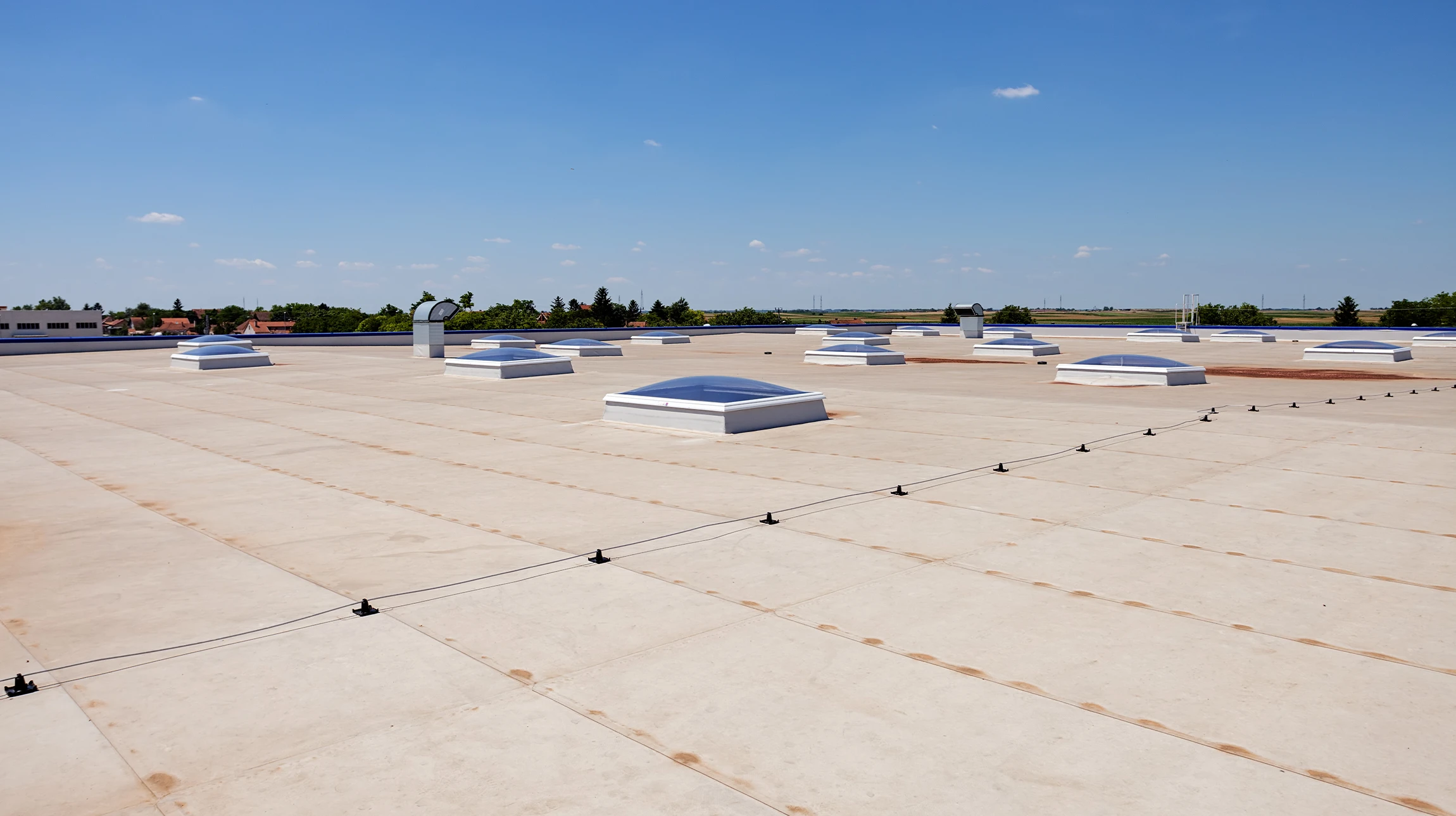 A flat commercial building roof with several skylights and rooftop vents, bordered by a safety cable, under a clear blue sky with a few small clouds. Trees and buildings are visible in the distance.