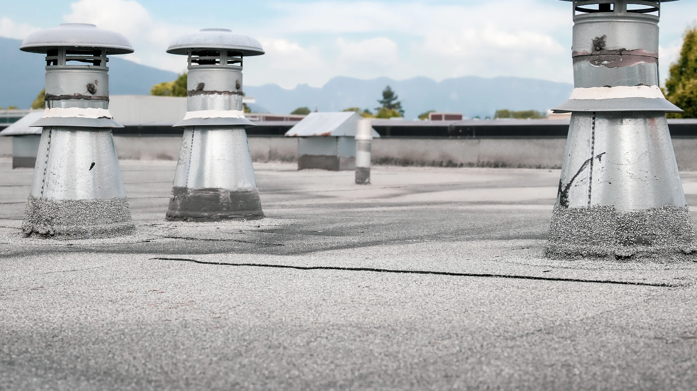 Three metal rooftop vents are positioned on a flat, gray roof with visible cracks. In the background, there are mountains, trees, and a partly cloudy sky.