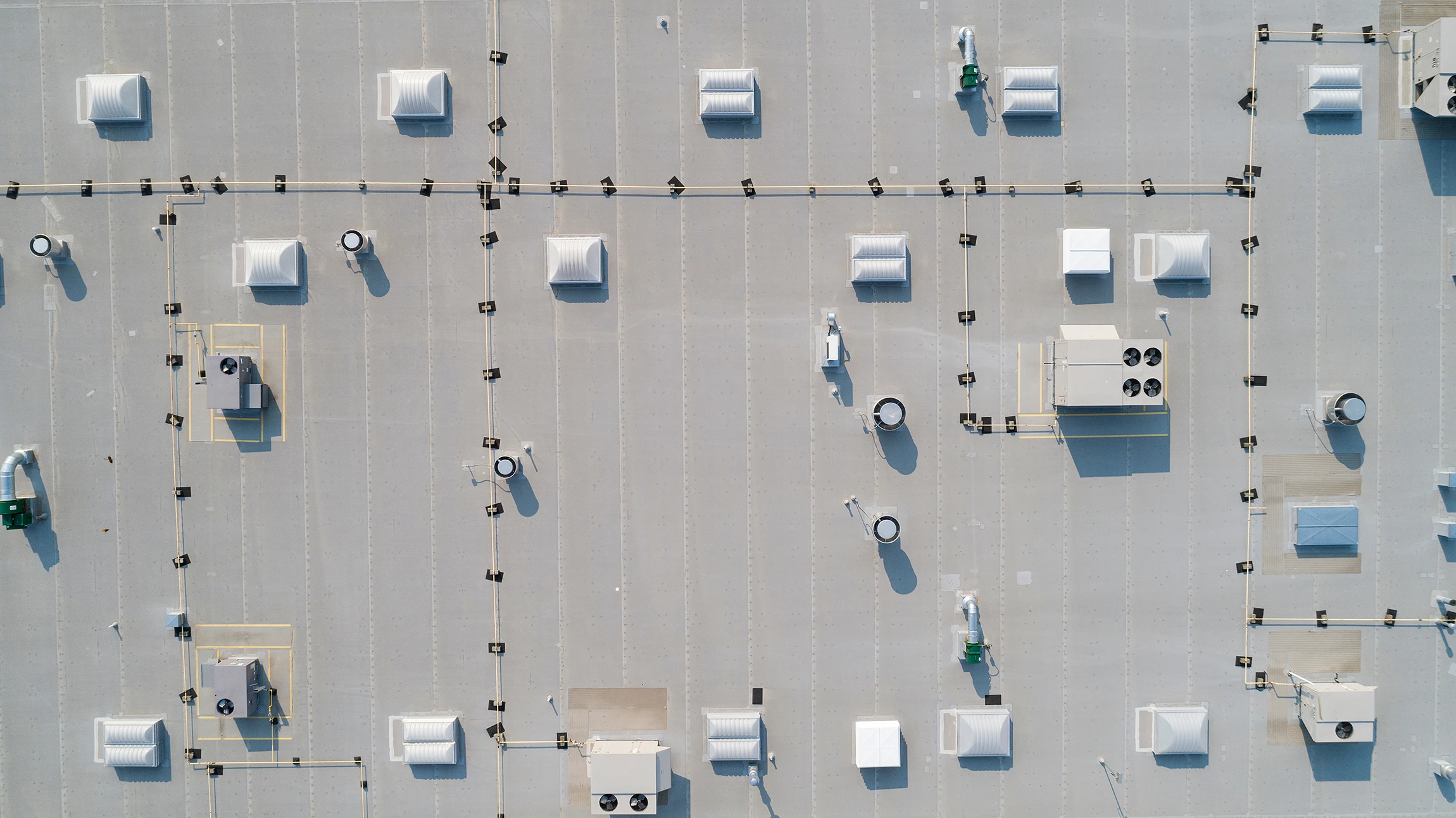 Aerial view of a large, flat industrial rooftop with numerous vents, HVAC units, and machinery evenly spaced across the light gray surface. Thin lines and small posts or barriers create a grid pattern.