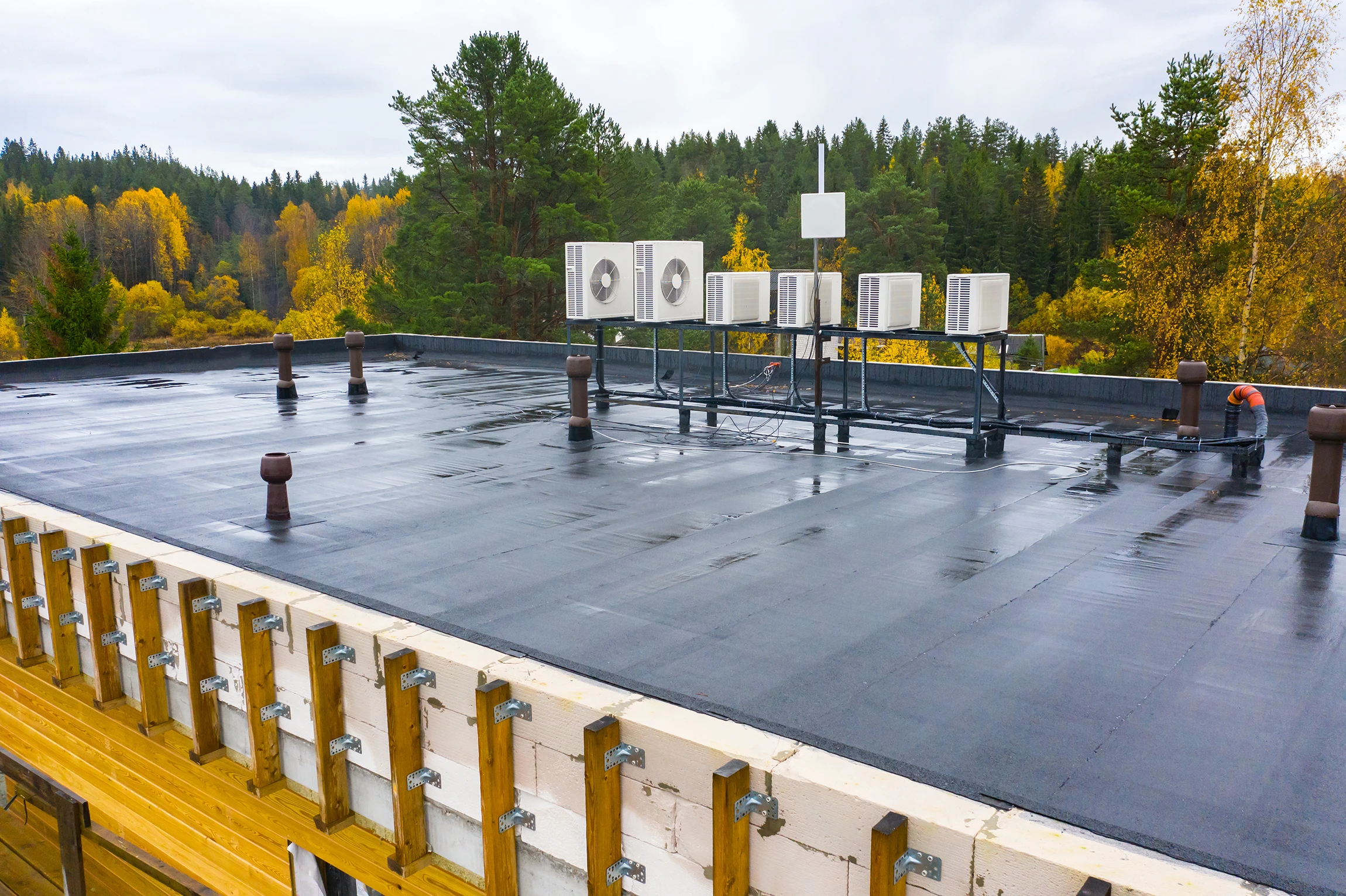 A flat rooftop covered with black waterproofing material features multiple air conditioning units on metal stands; surrounding are trees with autumn foliage.