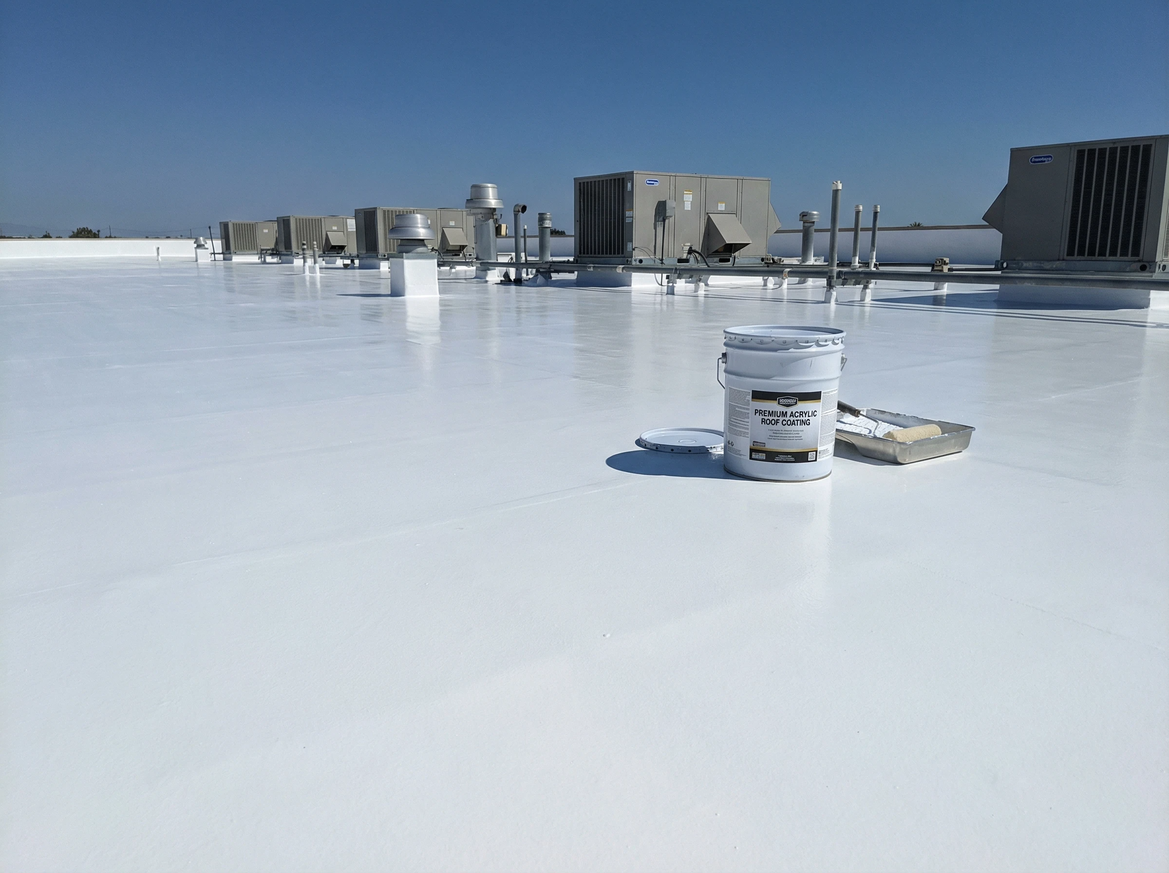 A bright white commercial rooftop with HVAC units and a bucket of roof coating beside a paint tray and roller, under a clear blue sky. The surface appears freshly coated and clean.