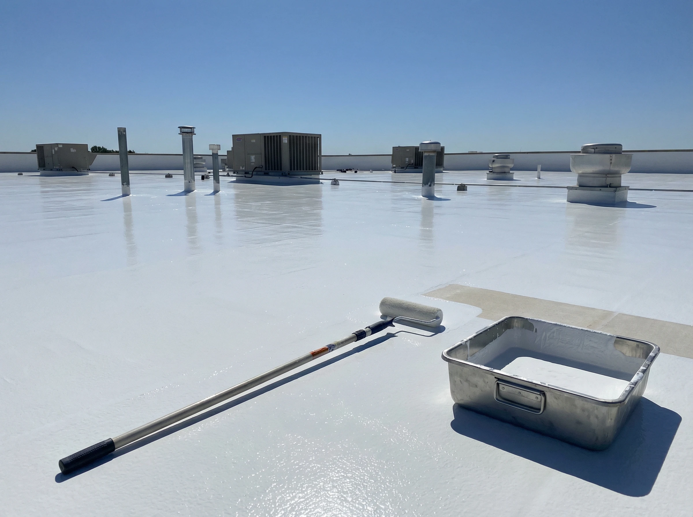 A rooftop coated in white sealant under a clear blue sky, with ventilation units and a paint roller and tray in the foreground, indicating ongoing roof maintenance work.