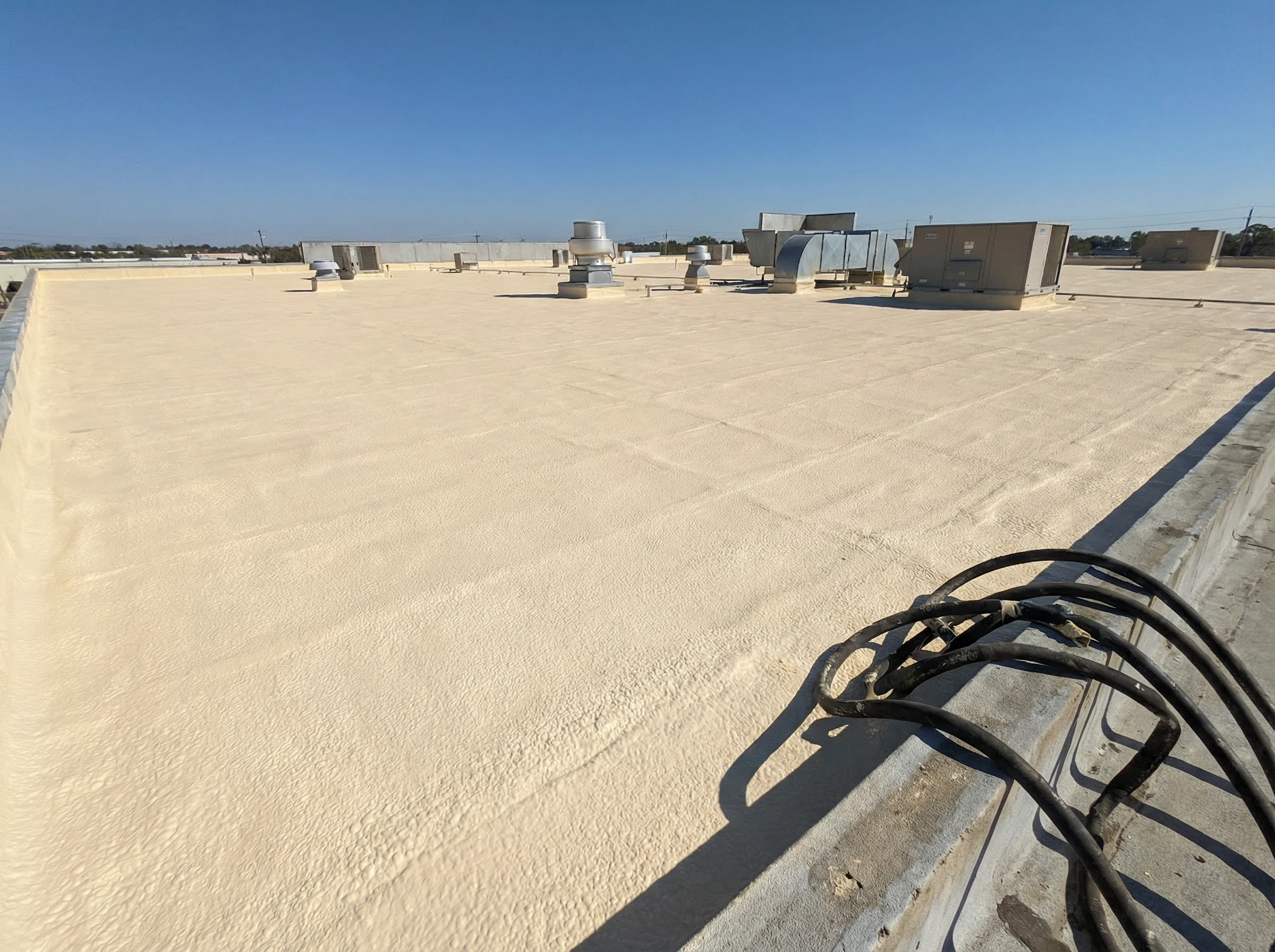 A flat commercial rooftop coated with a light-colored material, featuring various HVAC units and vents scattered across the surface under a clear blue sky. Coiled cables rest in the foreground near the roof’s edge.