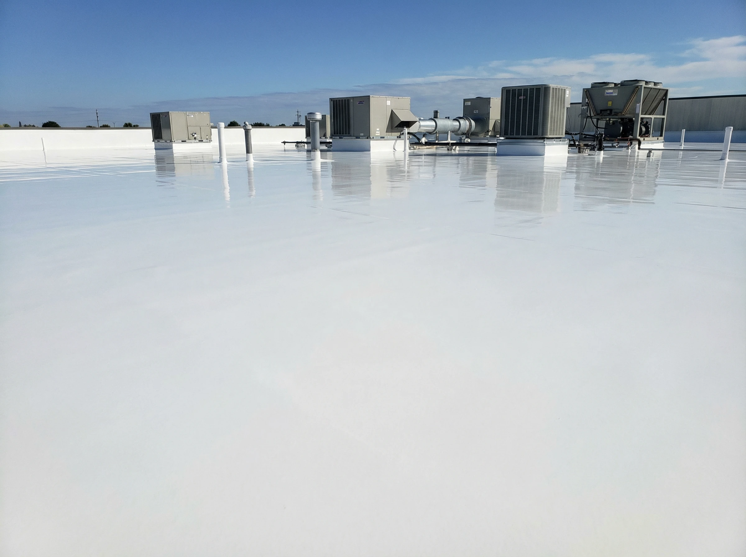 A flat, white commercial building rooftop with HVAC units and other equipment under a clear blue sky. The smooth, reflective roof surface extends across the image.