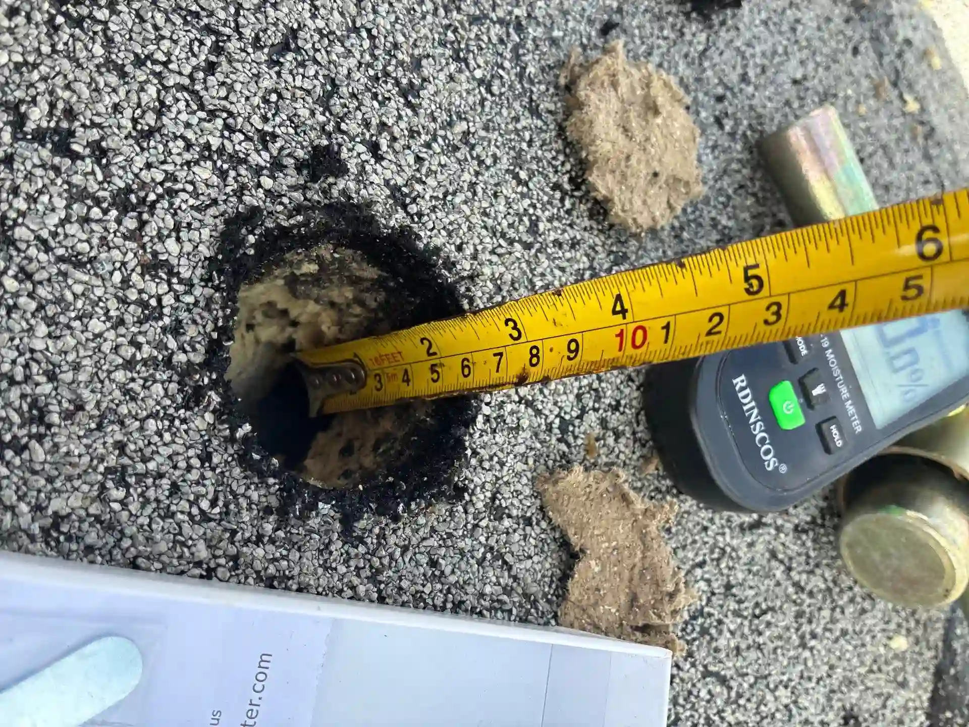 A yellow tape measure shows a hole in a rough, gray surface is about 4 inches wide. Nearby are insulation material, a pen, a digital device, and a piece of paper.