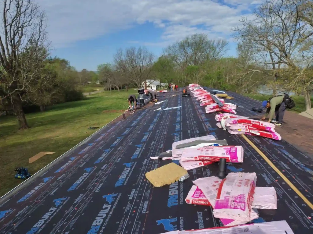 Workers are installing roofing materials on a large roof covered with underlayment, with bundles of shingles lined up. The surrounding area has green grass, trees, and a partly cloudy sky.