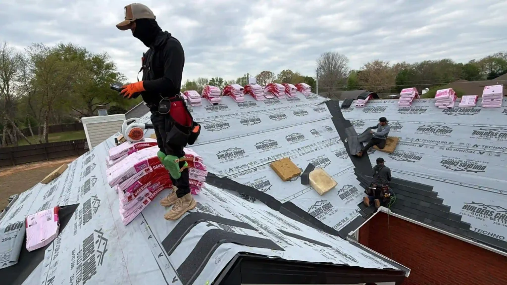 Workers install roofing materials on a house. One worker stands on the roof in protective clothing and gloves, while others work sitting or kneeling. Bundles of shingles are placed along the roof under a cloudy sky.
