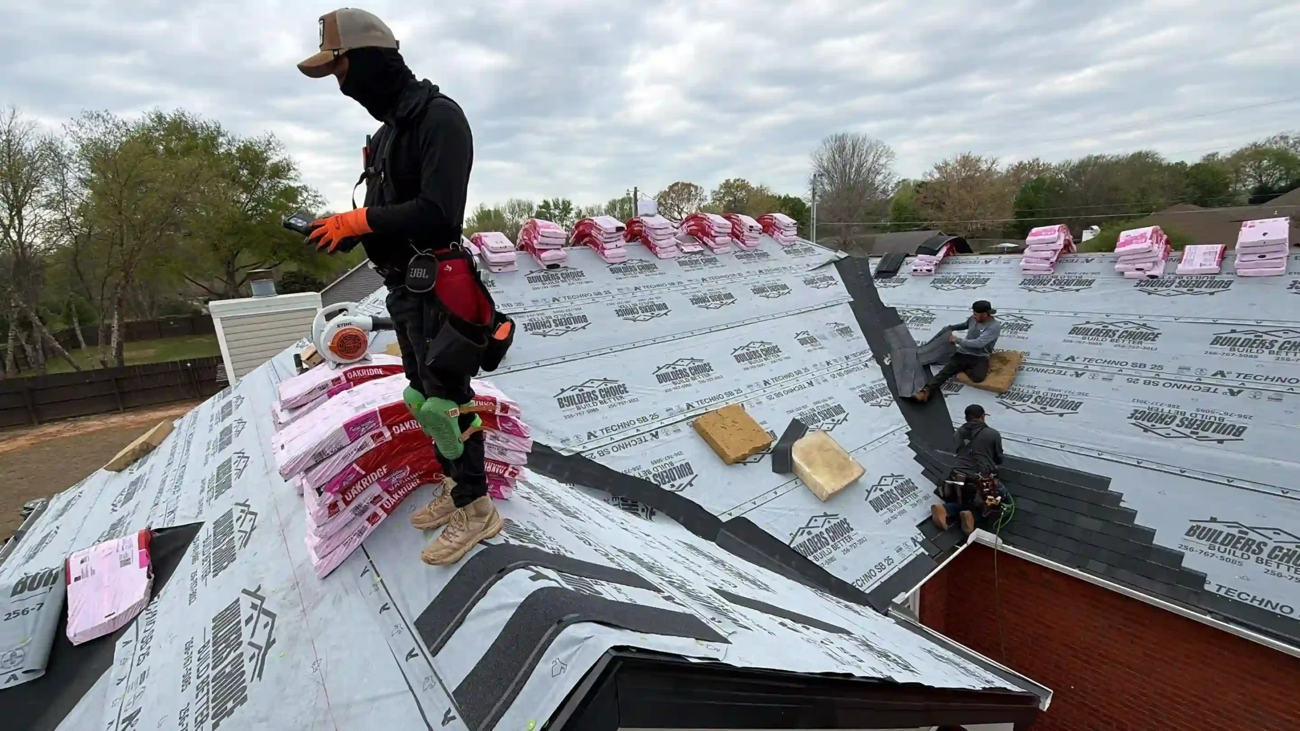 Workers install roofing materials on a house. One worker stands on the roof in protective clothing and gloves, while others work sitting or kneeling. Bundles of shingles are placed along the roof under a cloudy sky.