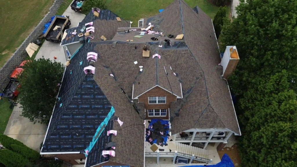 Aerial view of a large house with brown shingles, where part of the roof is being repaired. Roofing materials and workers are visible, and one section is covered with black underlayment. Tarps protect areas below.