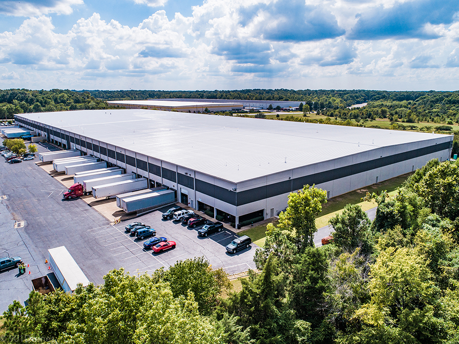 A large warehouse with a white roof, several loading docks, parked trucks, and cars. Surrounded by trees and greenery, with a partly cloudy sky overhead.