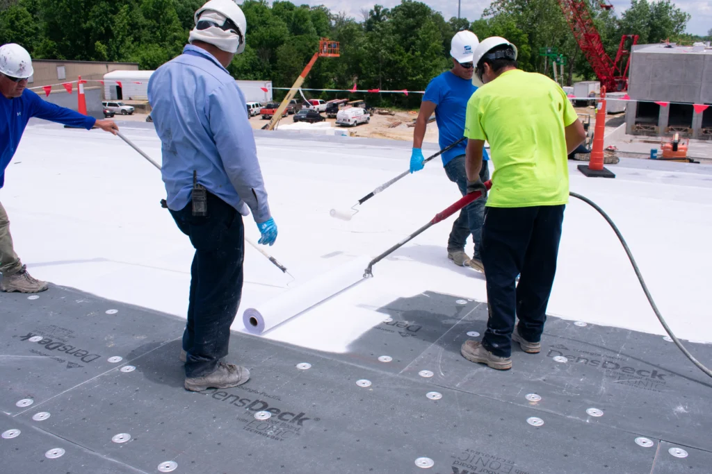 Four construction workers wearing helmets and safety gear are applying a white roofing material using rollers and a hose on a flat roof at a construction site surrounded by equipment and vehicles.