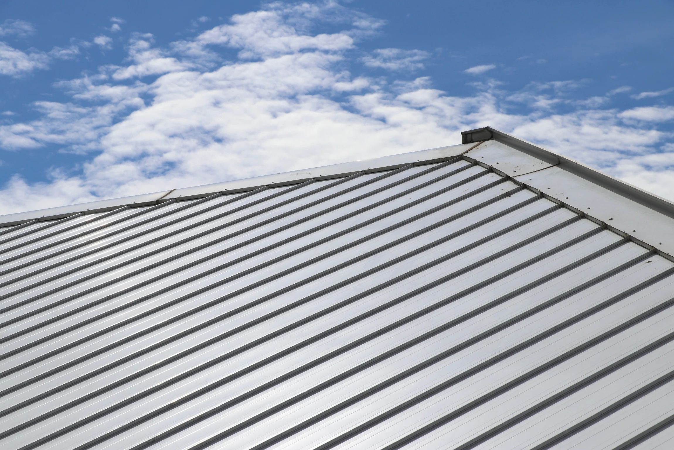A close-up view of a silver metal roof with vertical ridges, set against a blue sky with scattered white clouds.