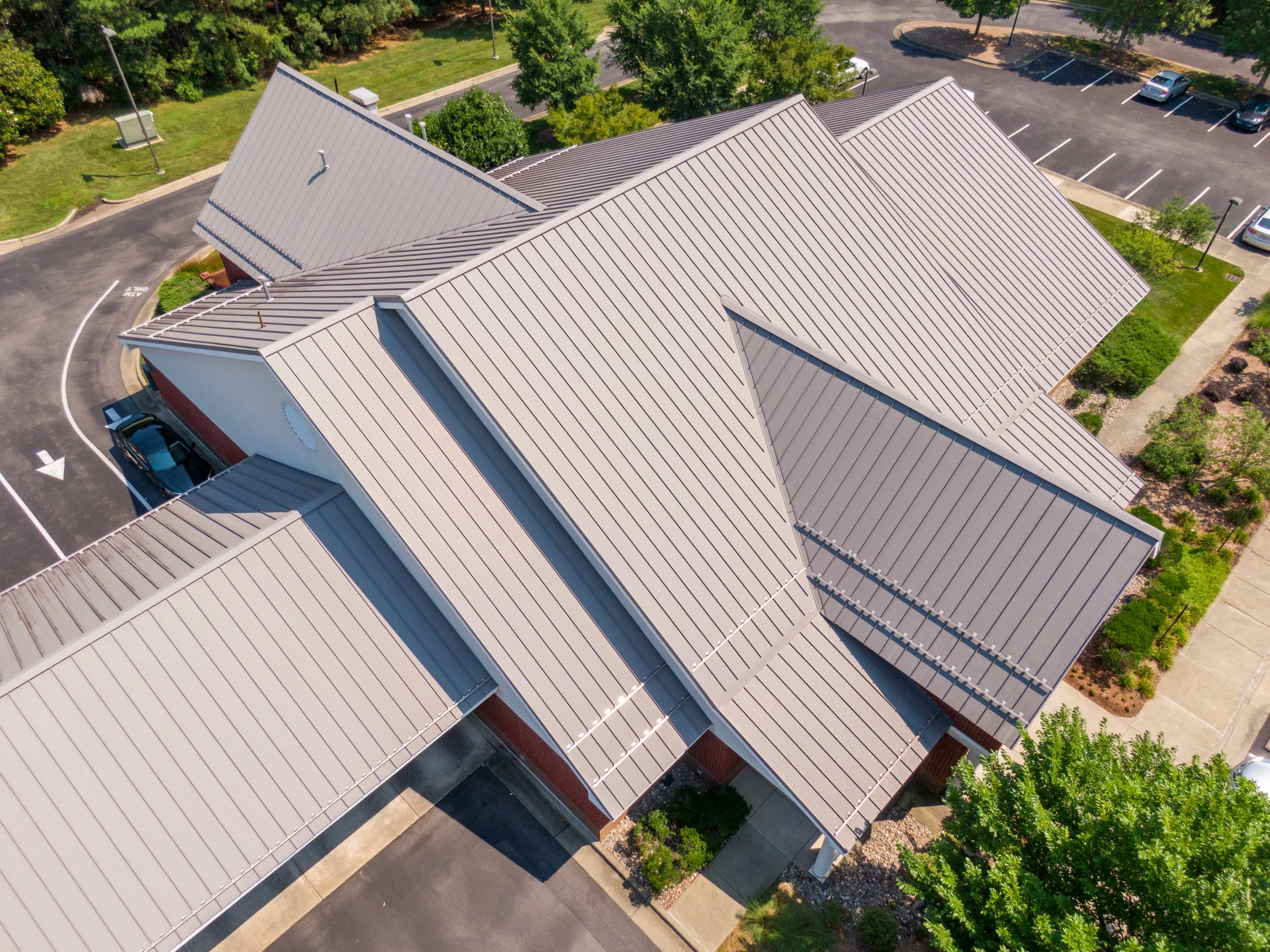 Aerial view of a building with a modern metal roof and multiple peaked sections, surrounded by trees, landscaped areas, and a mostly empty parking lot.