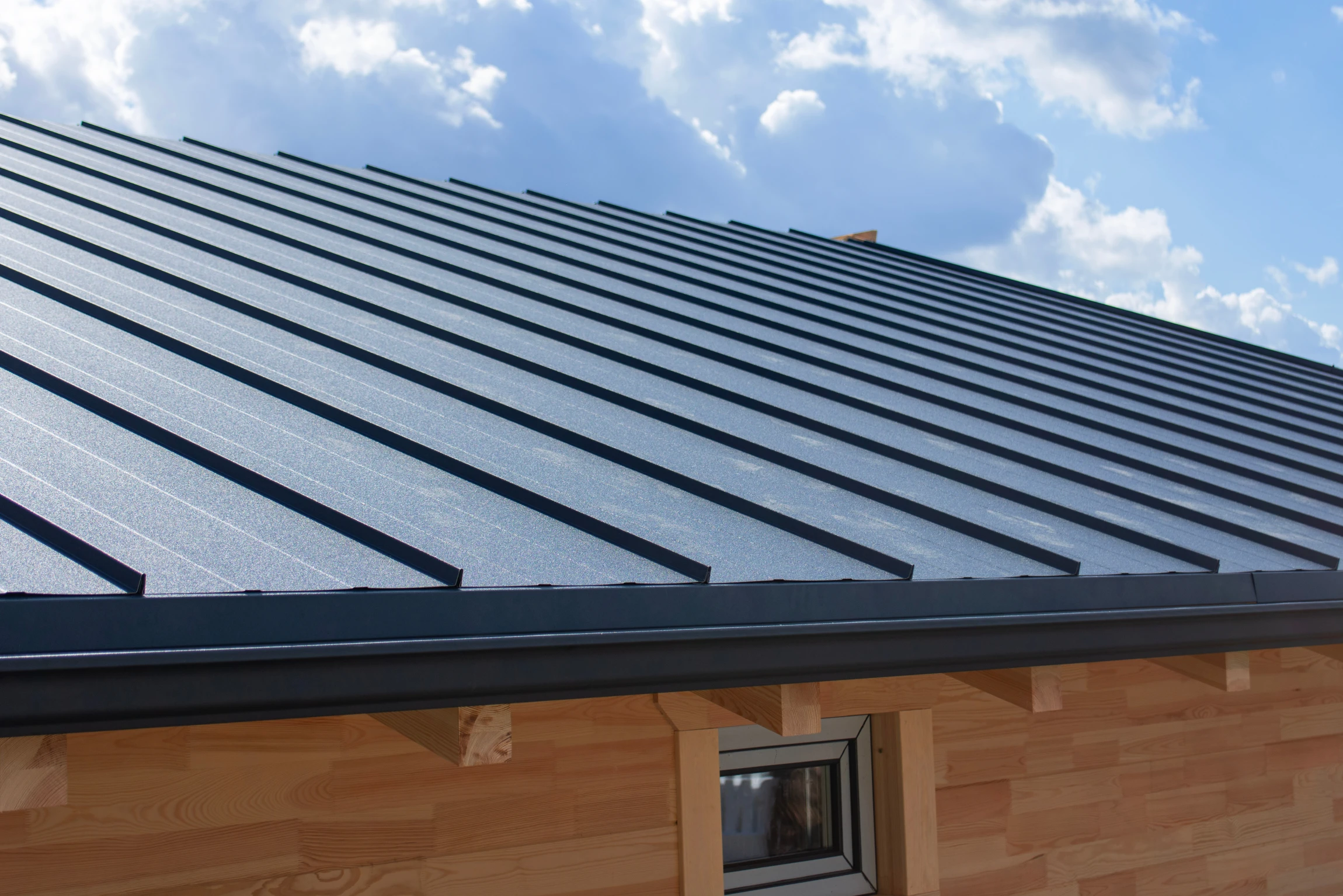 A close-up view of a modern metal roof on a wooden building, with a small window visible below the roof, set against a partly cloudy sky.