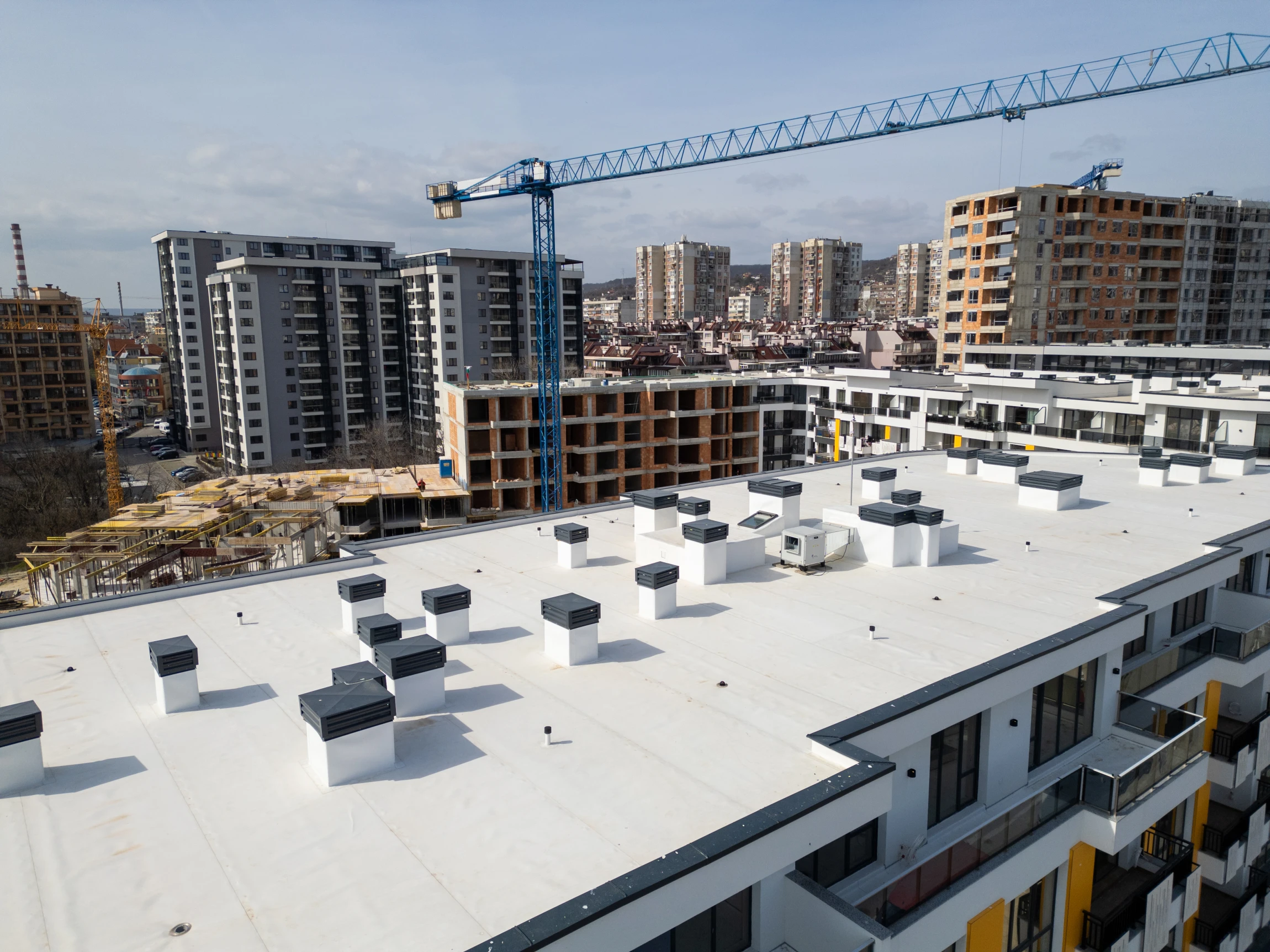Aerial view of modern apartment buildings under construction, with a blue crane in the background and completed buildings surrounding the site. Rooftop vents and balconies are visible in the foreground.