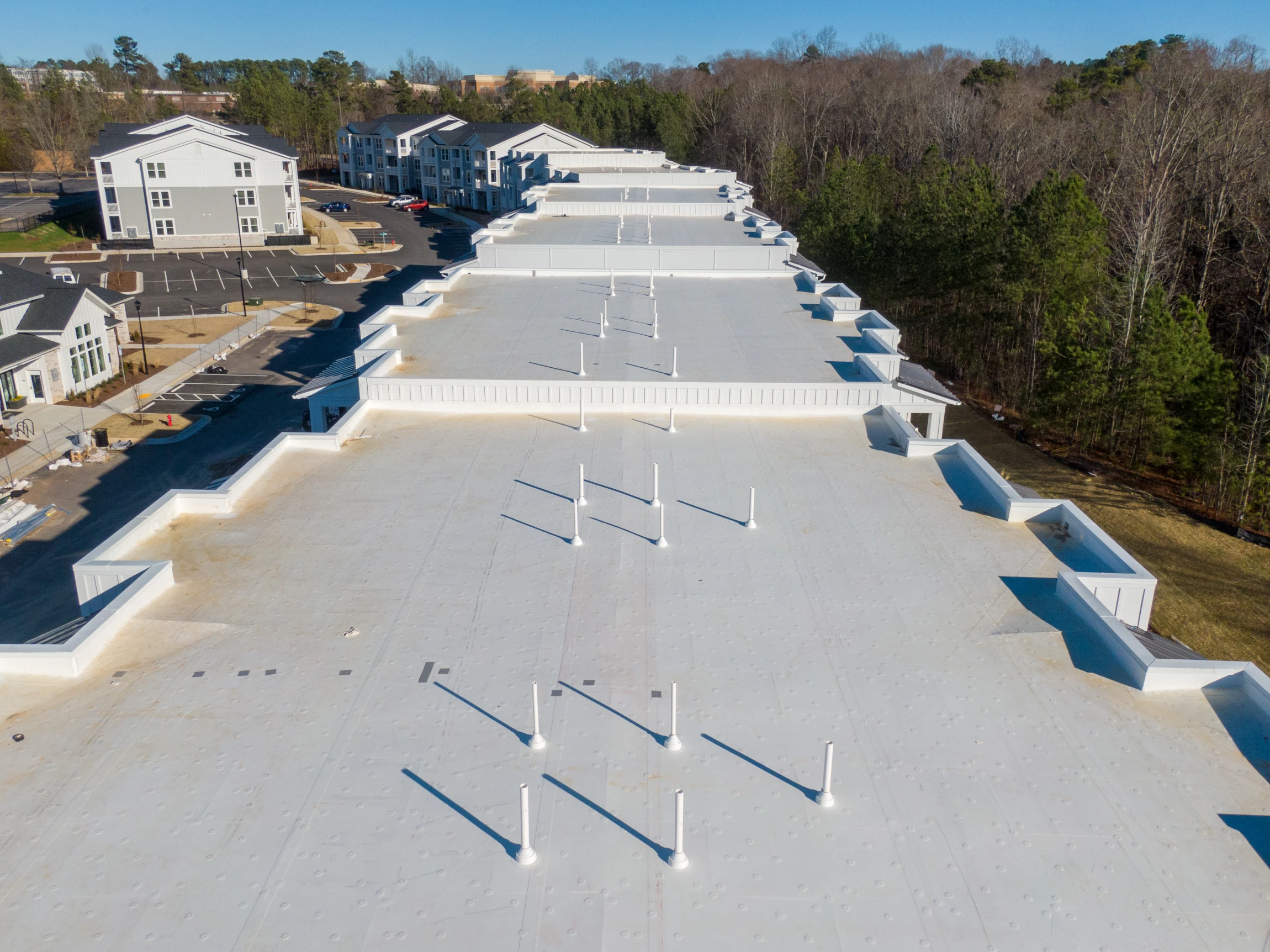 Aerial view of a large, flat white commercial roof with multiple vent pipes, surrounded by trees, parking lots, and nearby buildings on a clear day.