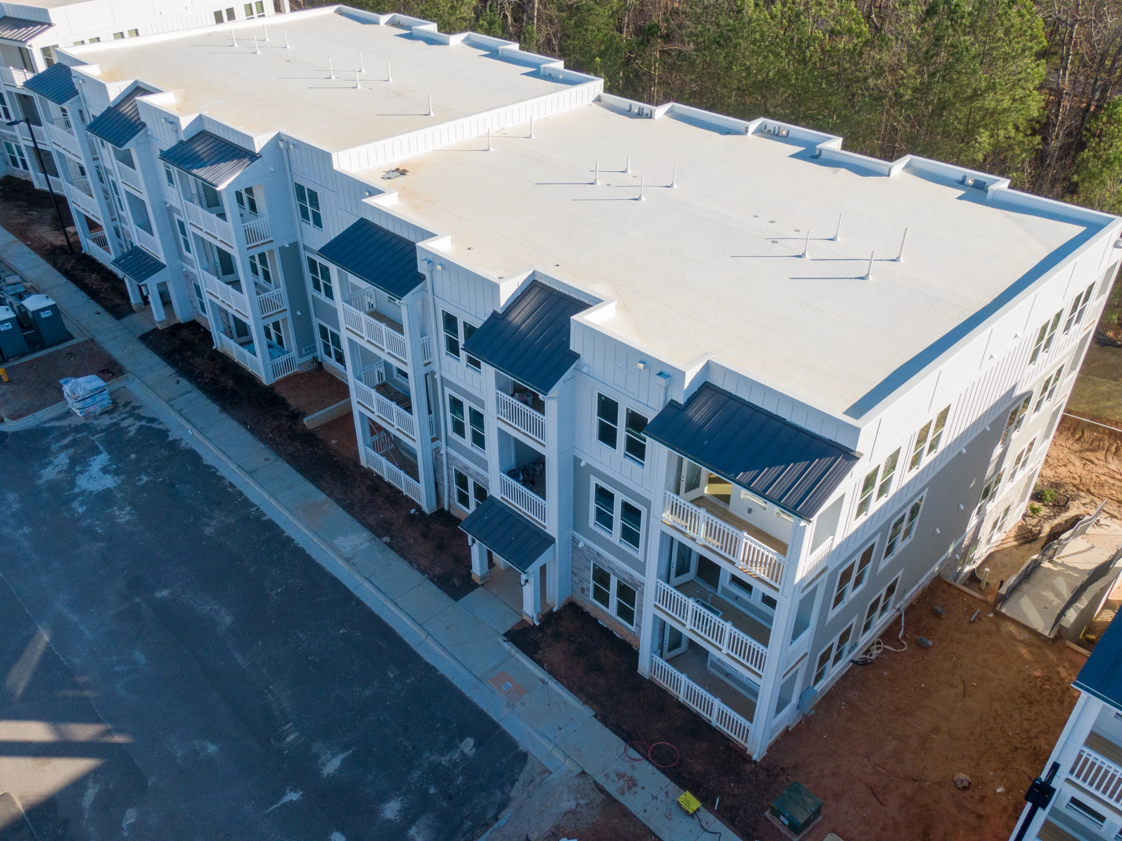 Aerial view of a modern, three-story apartment building with white siding, dark-colored roofs, multiple balconies, and surrounding construction areas next to a paved road and wooded area.