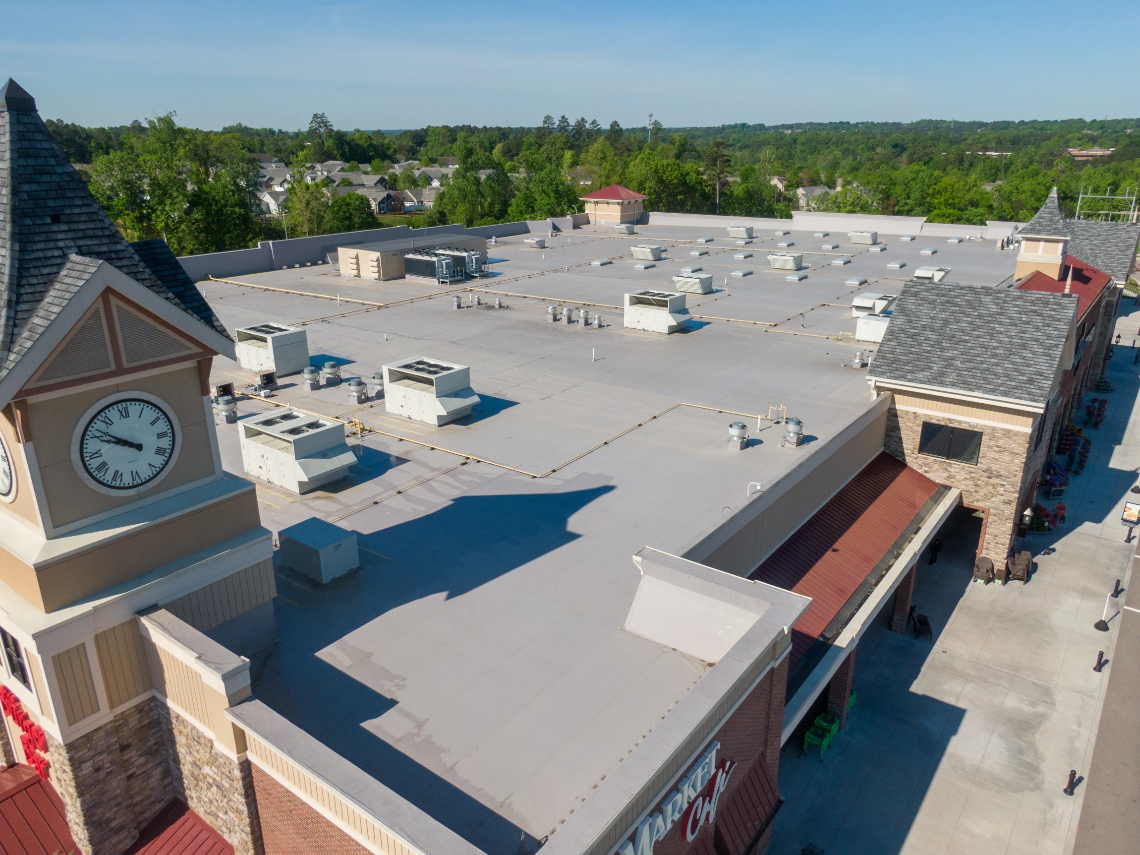 Aerial view of a shopping center with a large flat roof, several rooftop HVAC units, and a clock tower; surrounded by trees and other buildings on a sunny day.
