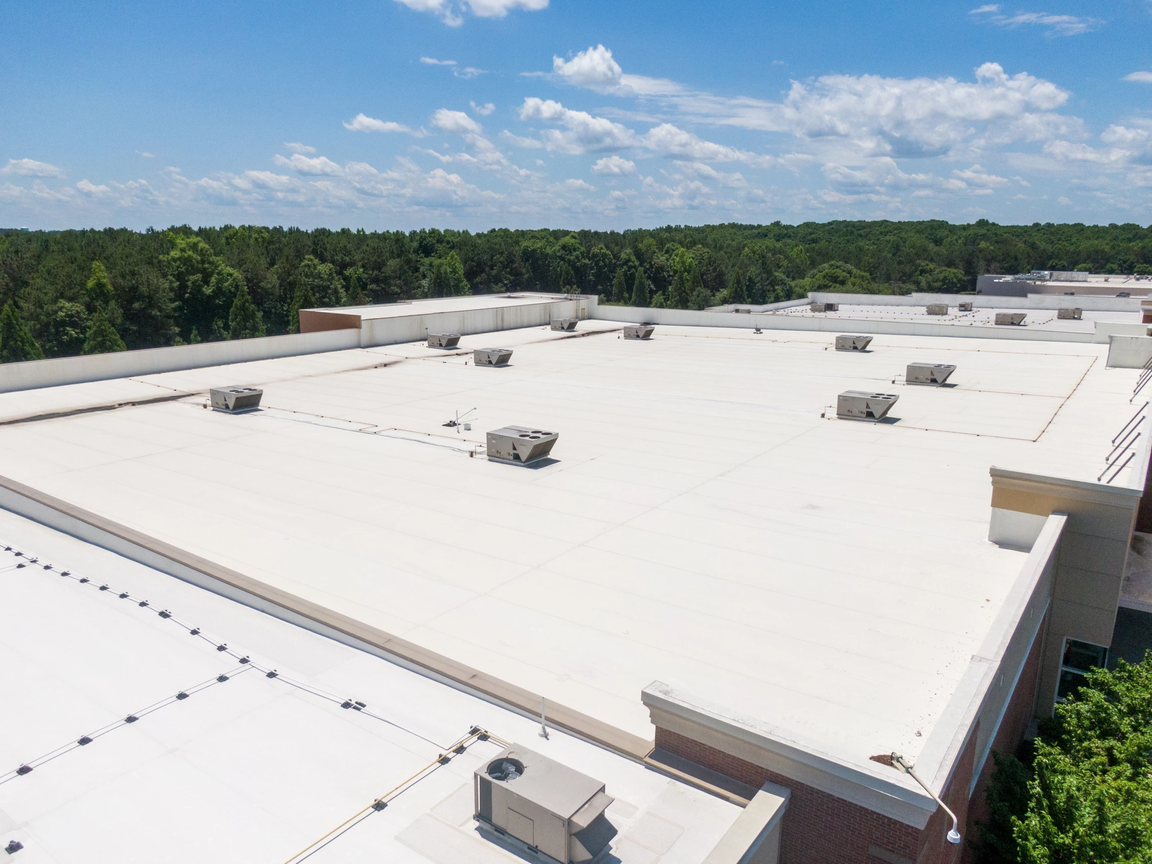 A large, flat white commercial rooftop with several HVAC units, surrounded by trees and a partly cloudy sky in the background.