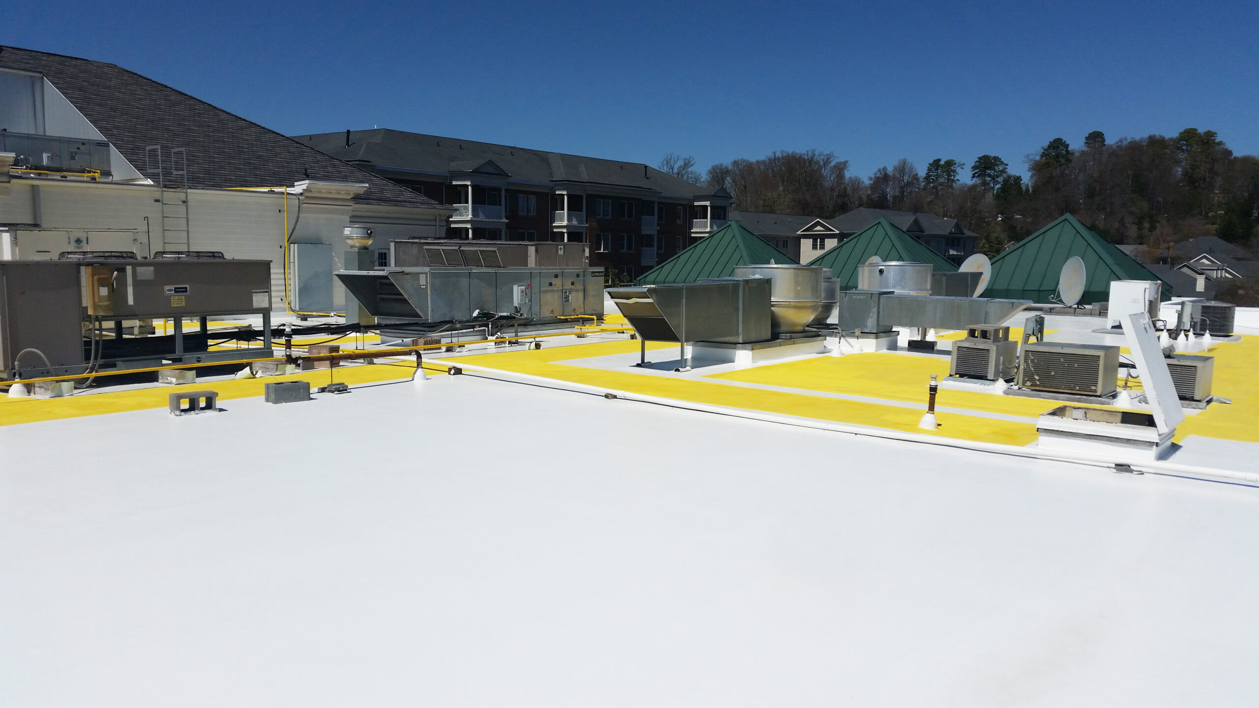 Flat commercial building roof with white and yellow reflective coating, featuring multiple HVAC units and ventilation systems, under a clear blue sky with residential buildings and trees in the background.