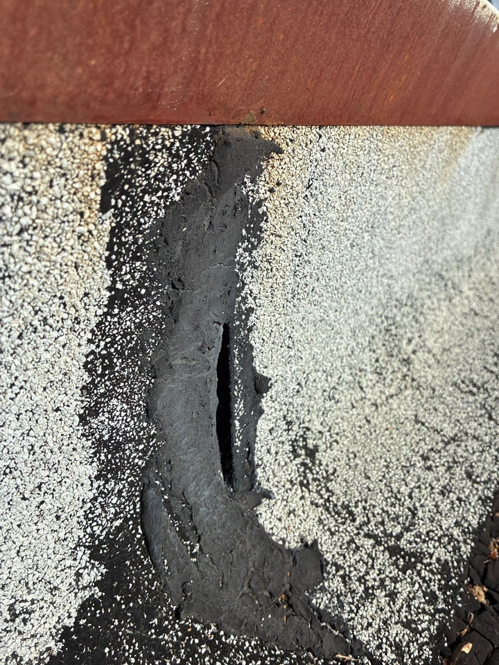 Close-up of a damaged section of a shingled roof with a vertical tear or crack in the surface, exposing the darker layer beneath the shingles. A rust-colored edge runs along the top of the image.