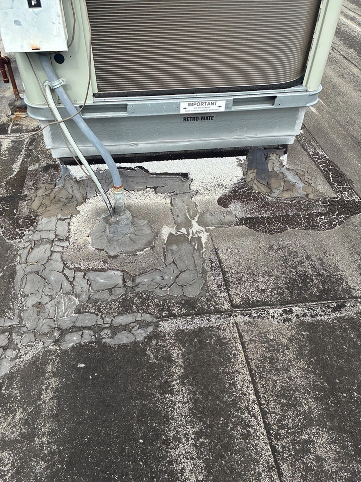 A rooftop HVAC unit with cracked, uneven, and patched sealant around its base, showing wear and weathering on the surrounding roofing surface.
