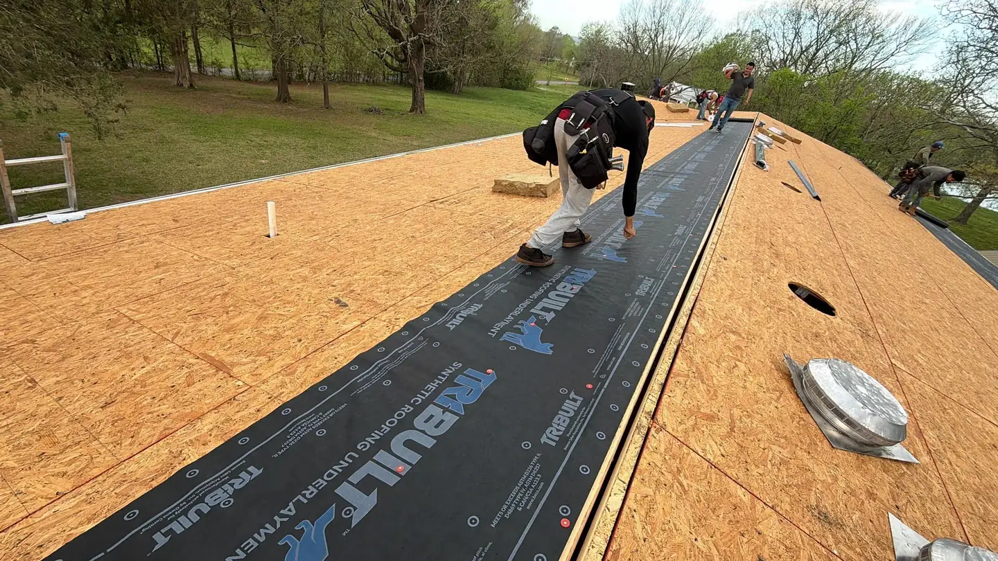 A worker in safety gear installs roofing underlayment on a sloped wooden roof while others work in the background; tools and roofing materials are scattered around, and trees and grass are visible beyond the roof.