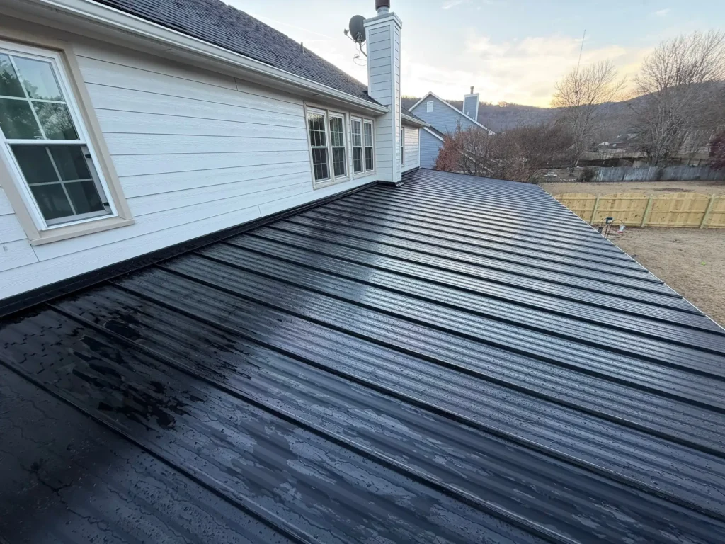 A black standing seam metal roof on a white house, viewed at an angle, wet with water streaks, with a fenced backyard behind.