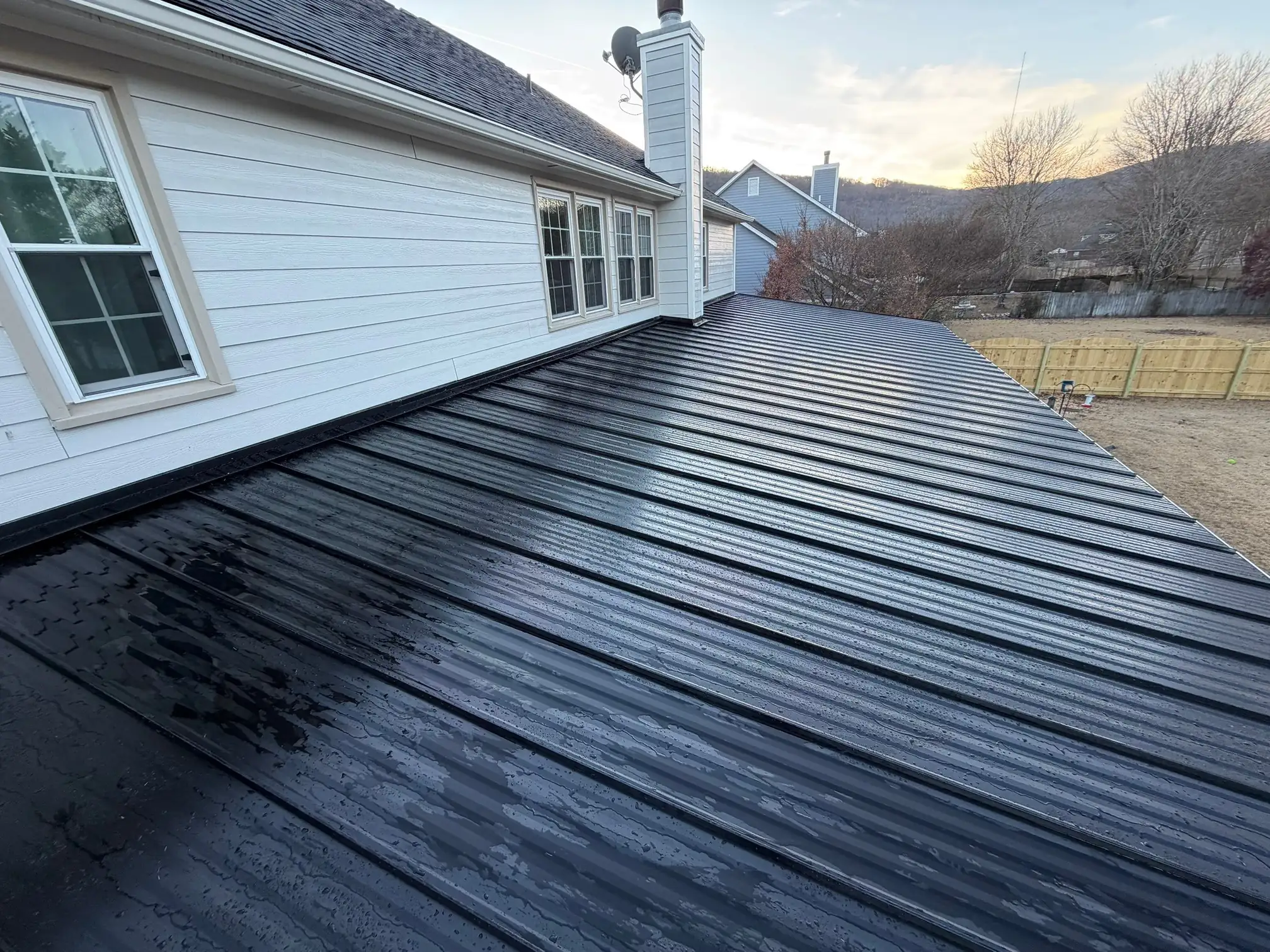 A black metal roof attached to a white house, viewed at an angle. The roof is wet with water streaks, and a fenced backyard and leafless trees are visible in the background.