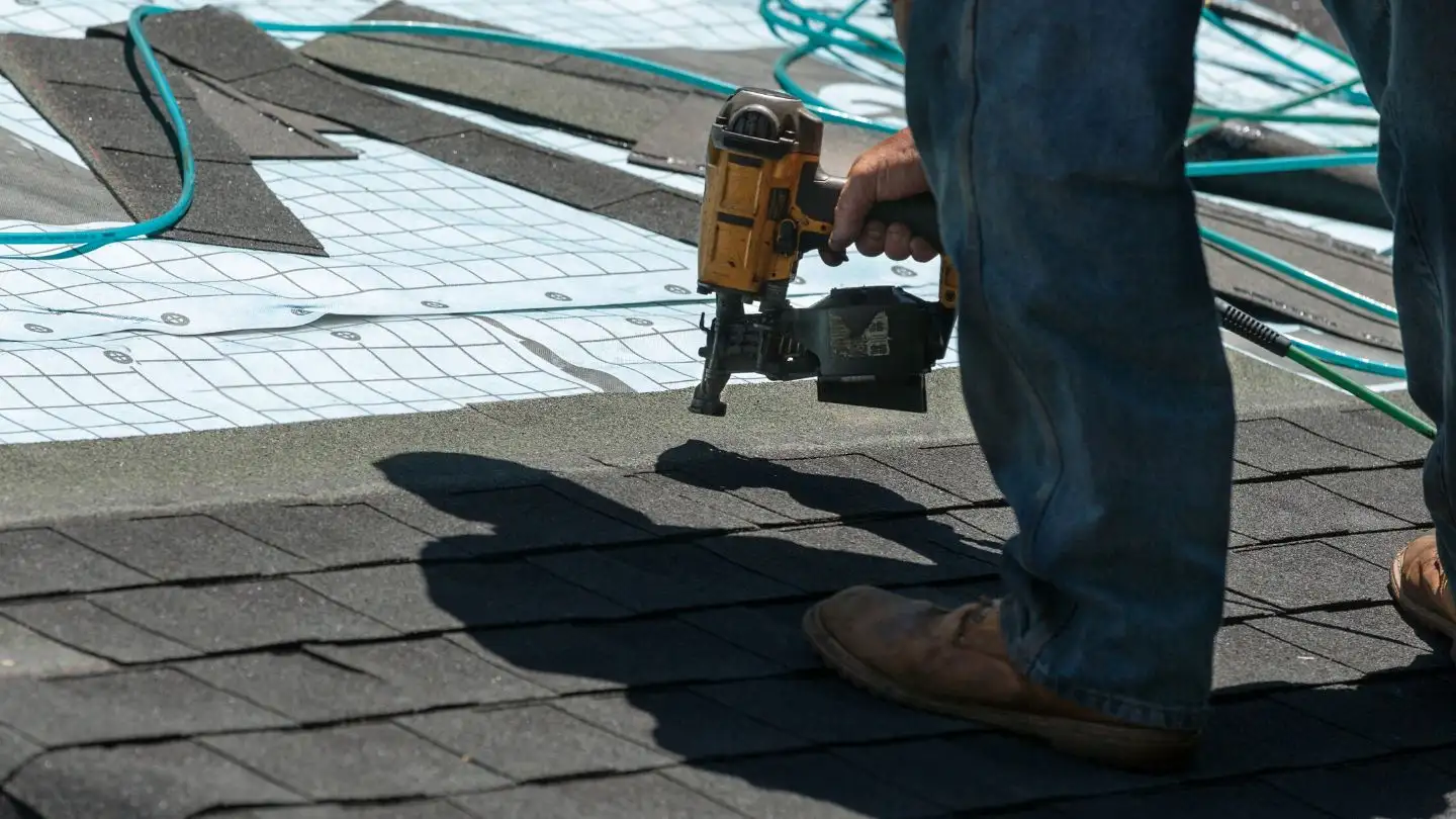 A person wearing jeans and boots uses a nail gun to install asphalt shingles on a roof, with roofing materials and a hose visible in the background.