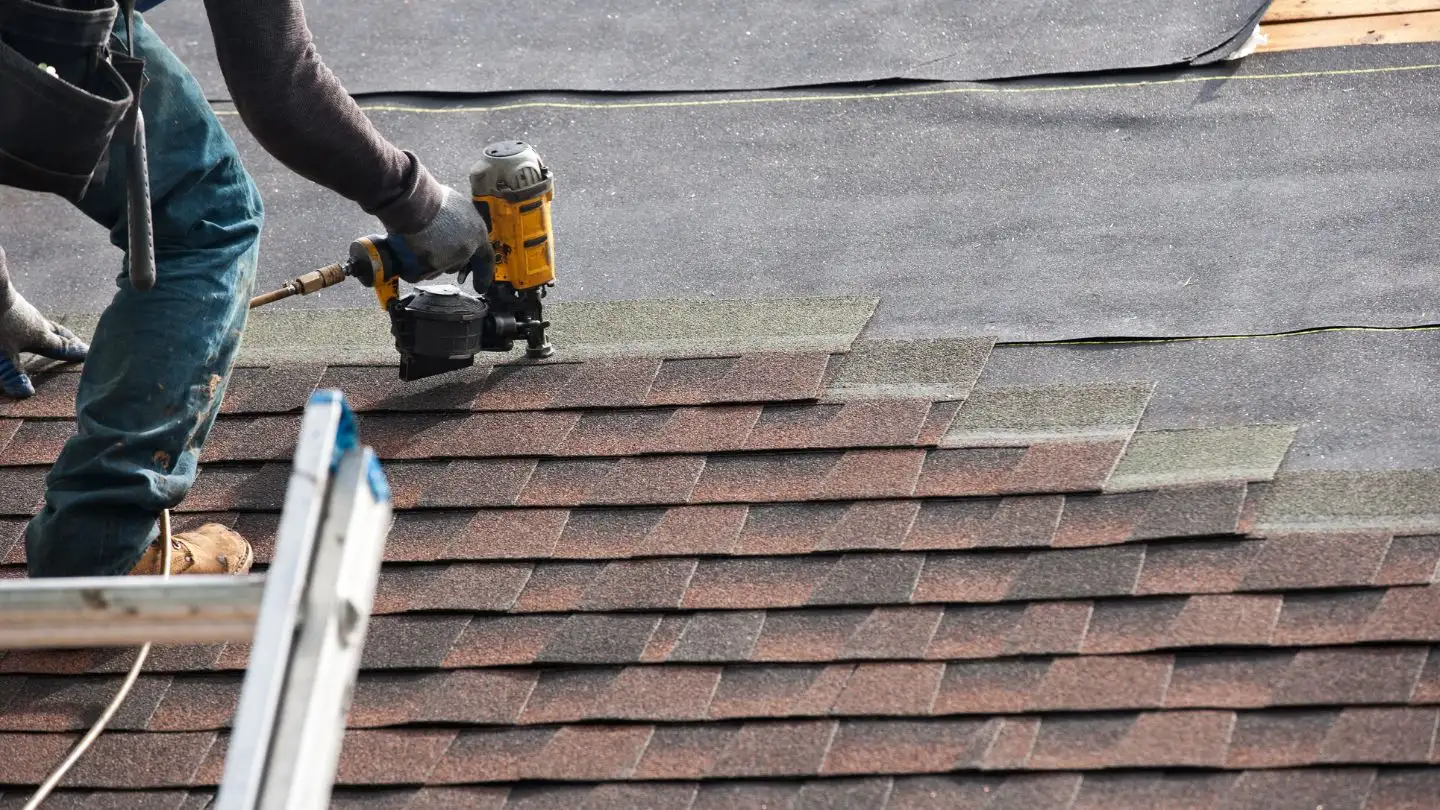 A worker uses a nail gun to install asphalt shingles on a roof. A ladder is visible, and rolls of roofing material are in the background.