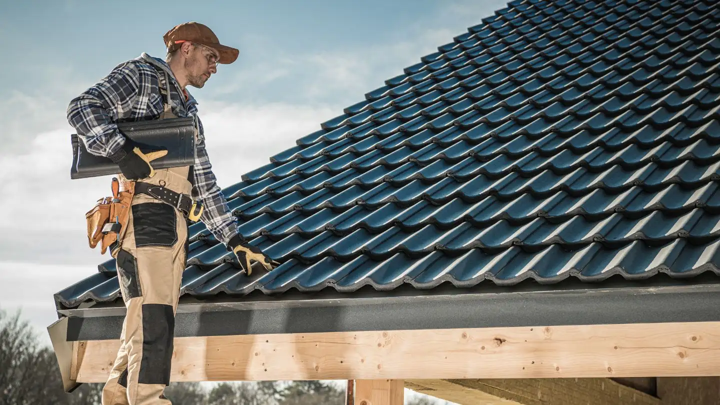 A construction worker in a plaid shirt and cap inspects a tiled roof while holding a folder, standing near the edge of a new wooden building structure.